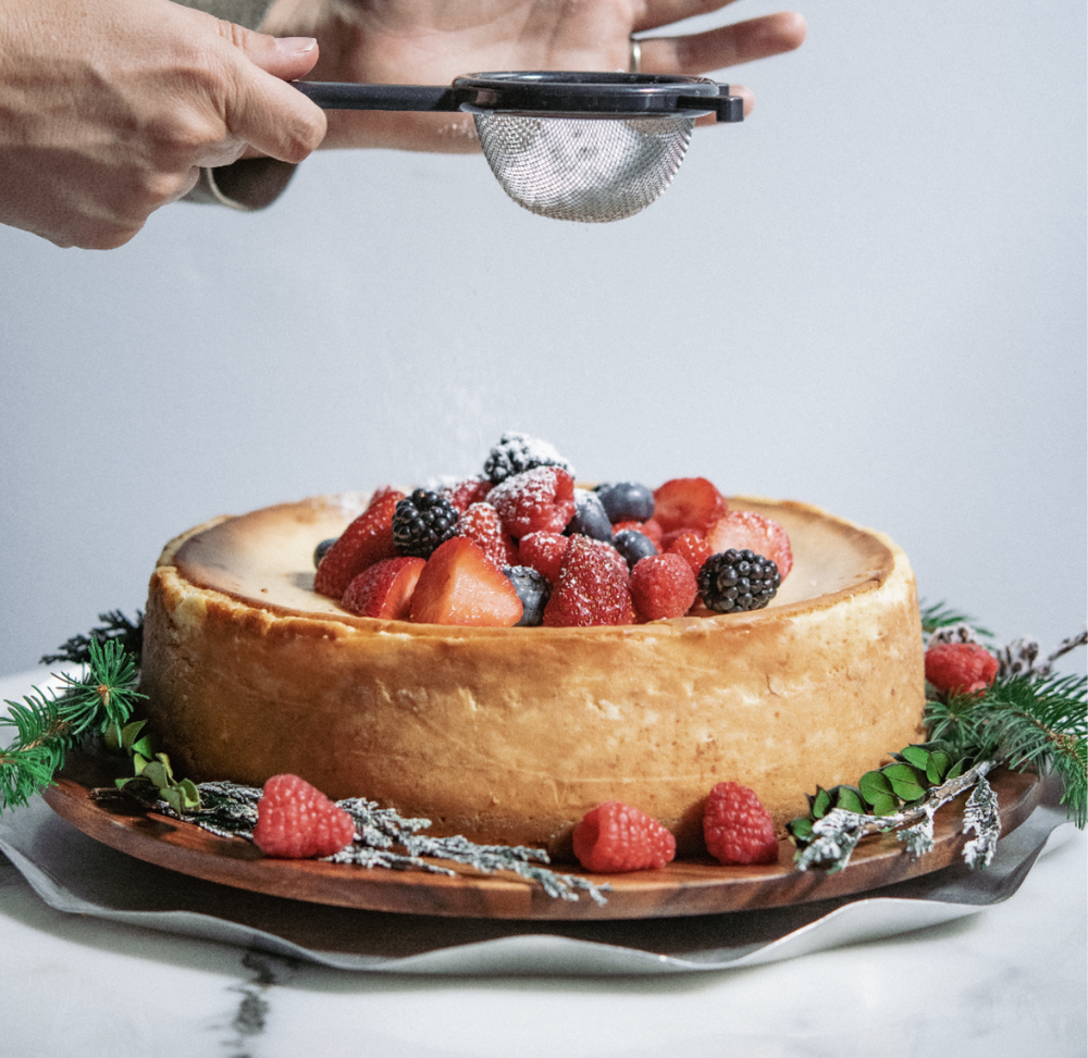 Cheesecake with berries being dusted with powdered sugar.