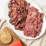 A plate with slices of pastrami and corned beef, with a small bowl of mustard and a flag marker with the Carnegie brand logo.