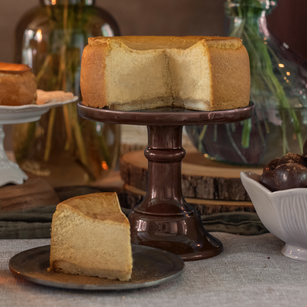 A pumpkin spice cheesecake on a brown cake stand with one slice served on a plate, alongside a dessert bowl and decorative elements in the background.